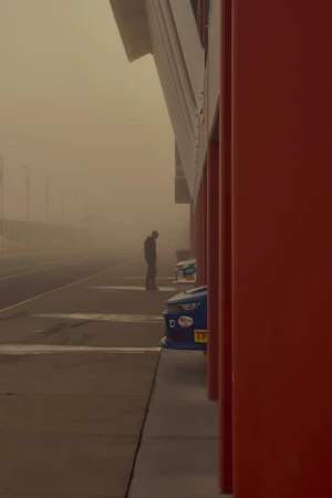 A man stands next to race cars in a garage on a foggy day. The row of pillars and the cars are vividly colorful and distinct.
