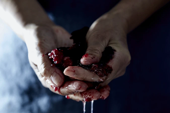 Close-up of a pair of hands holding crushed berries, juice dripping. The skin tone is fair, with a dark background.