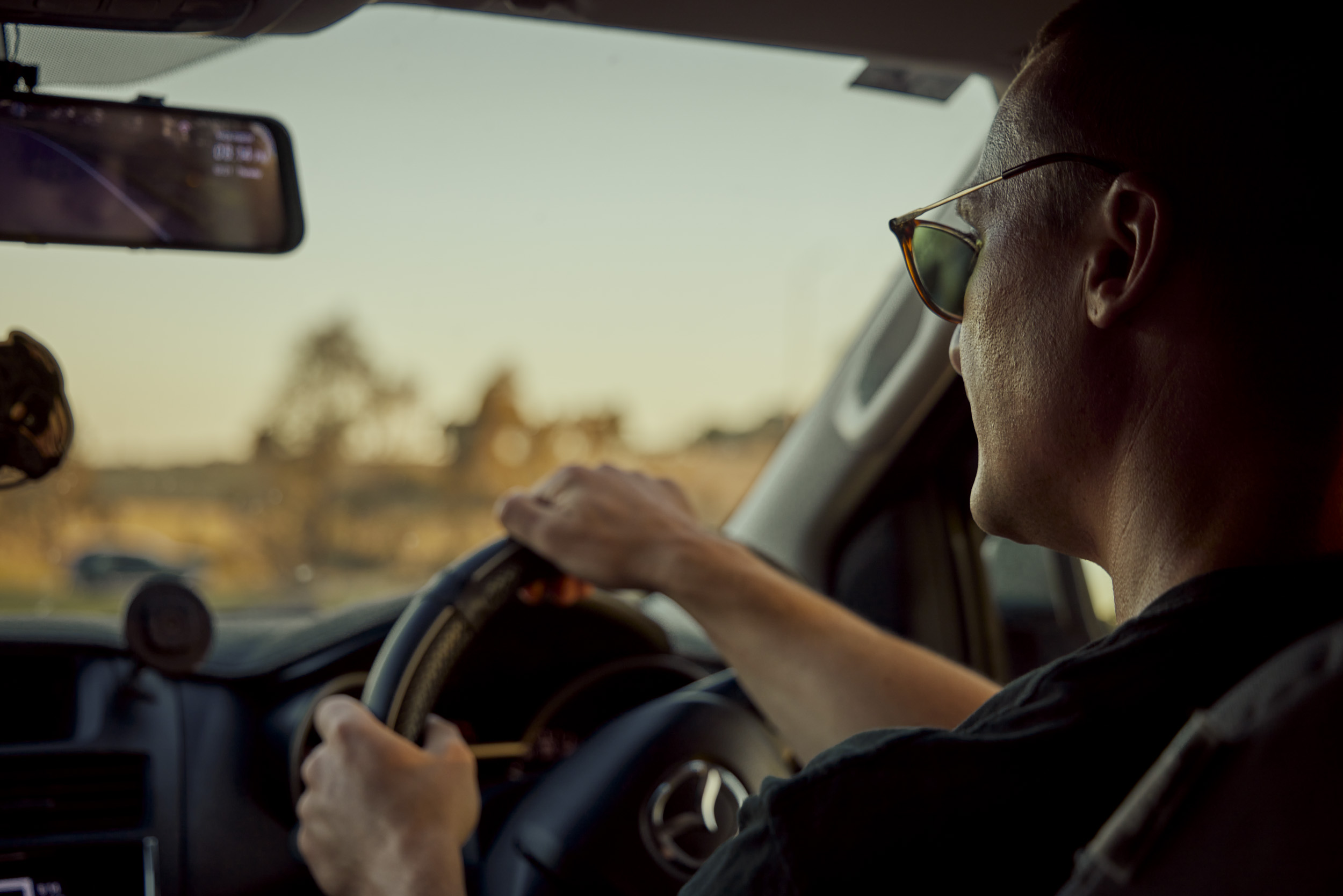 Man drives a car at dusk, wearing sunglasses, hands on steering wheel, visible dashboard and road ahead through windshield.