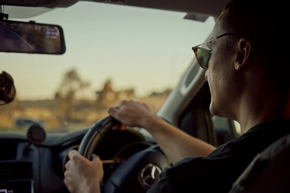 Man drives a car at dusk, wearing sunglasses, hands on steering wheel, visible dashboard and road ahead through windshield.