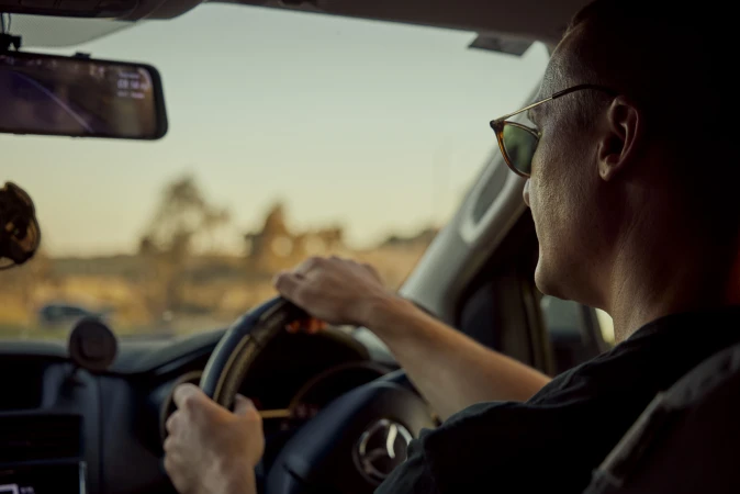 Man drives a car at dusk, wearing sunglasses, hands on steering wheel, visible dashboard and road ahead through windshield.