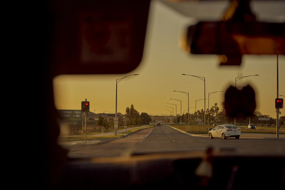 View through a car windshield of an empty highway at sunset with a sedan, street lights, and signage indicating nearby schools and parks.