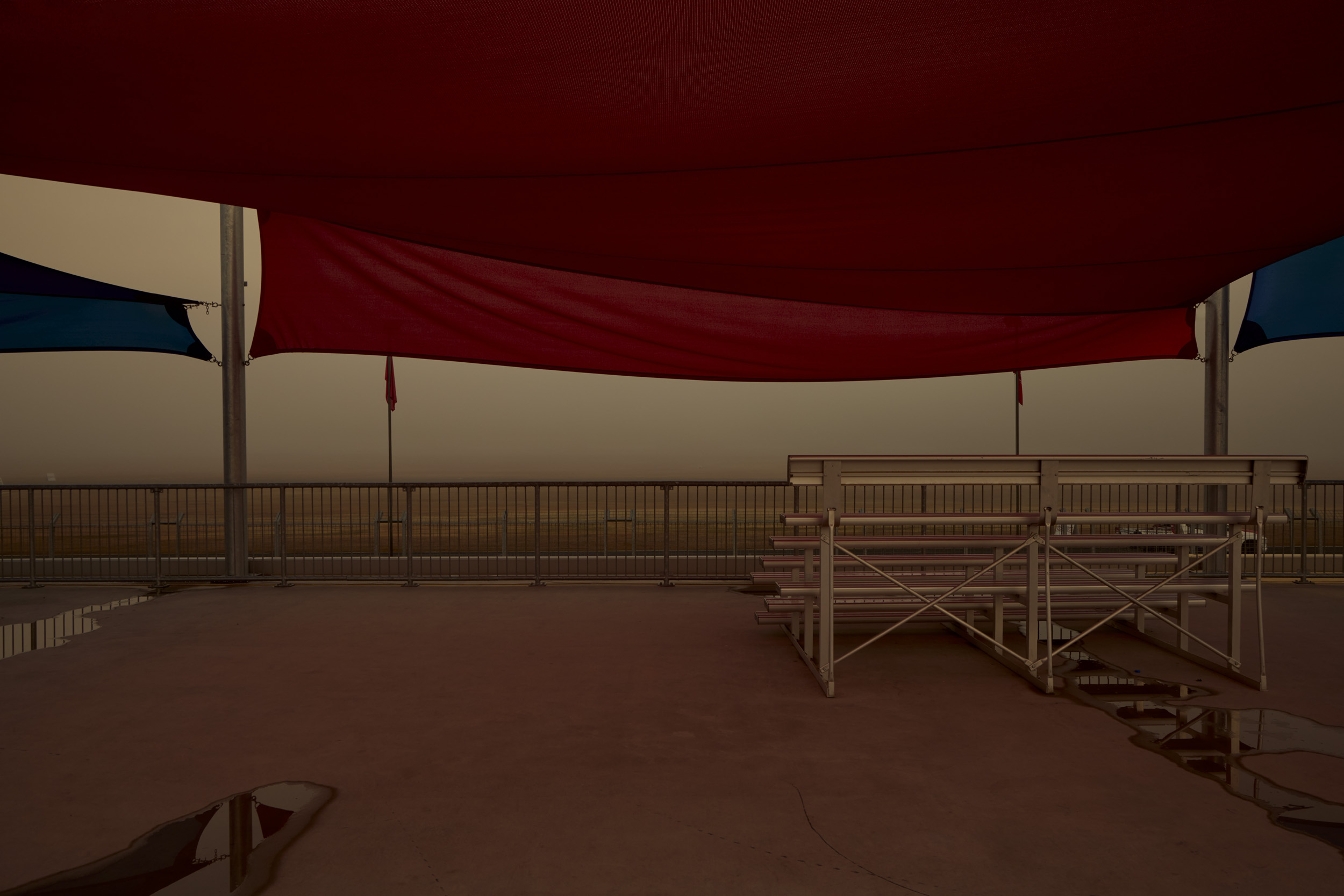 Empty metal bleachers sit on a concrete surface underneath red and blue shade structures with overcast sky background.