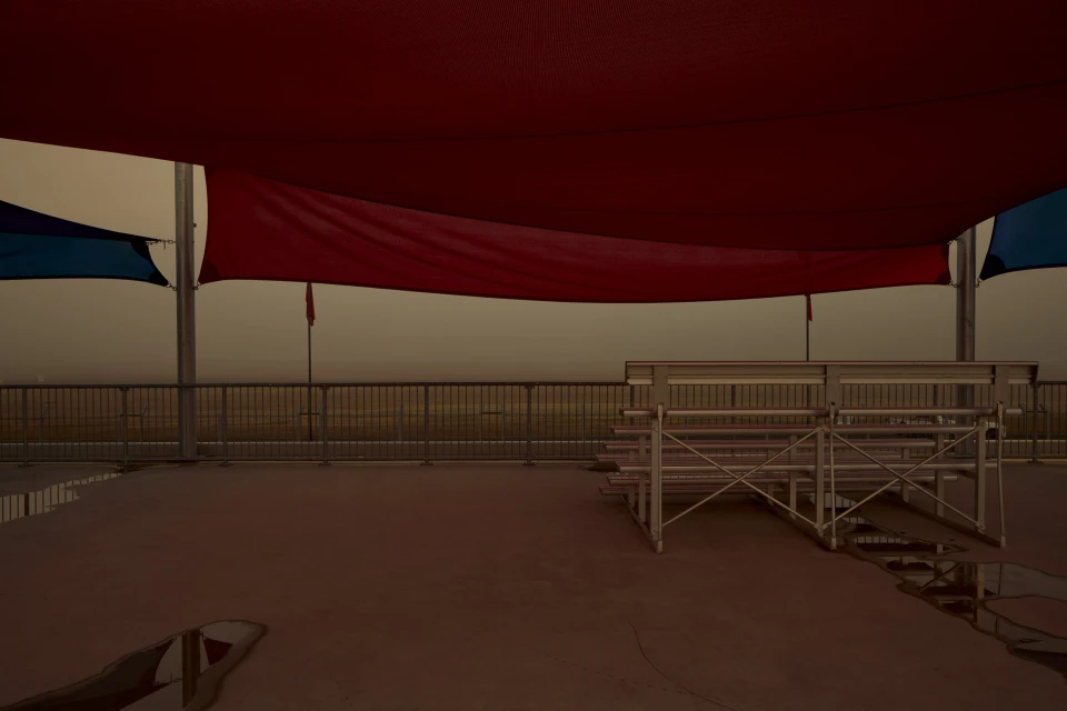 Empty metal bleachers sit on a concrete surface underneath red and blue shade structures with overcast sky background.