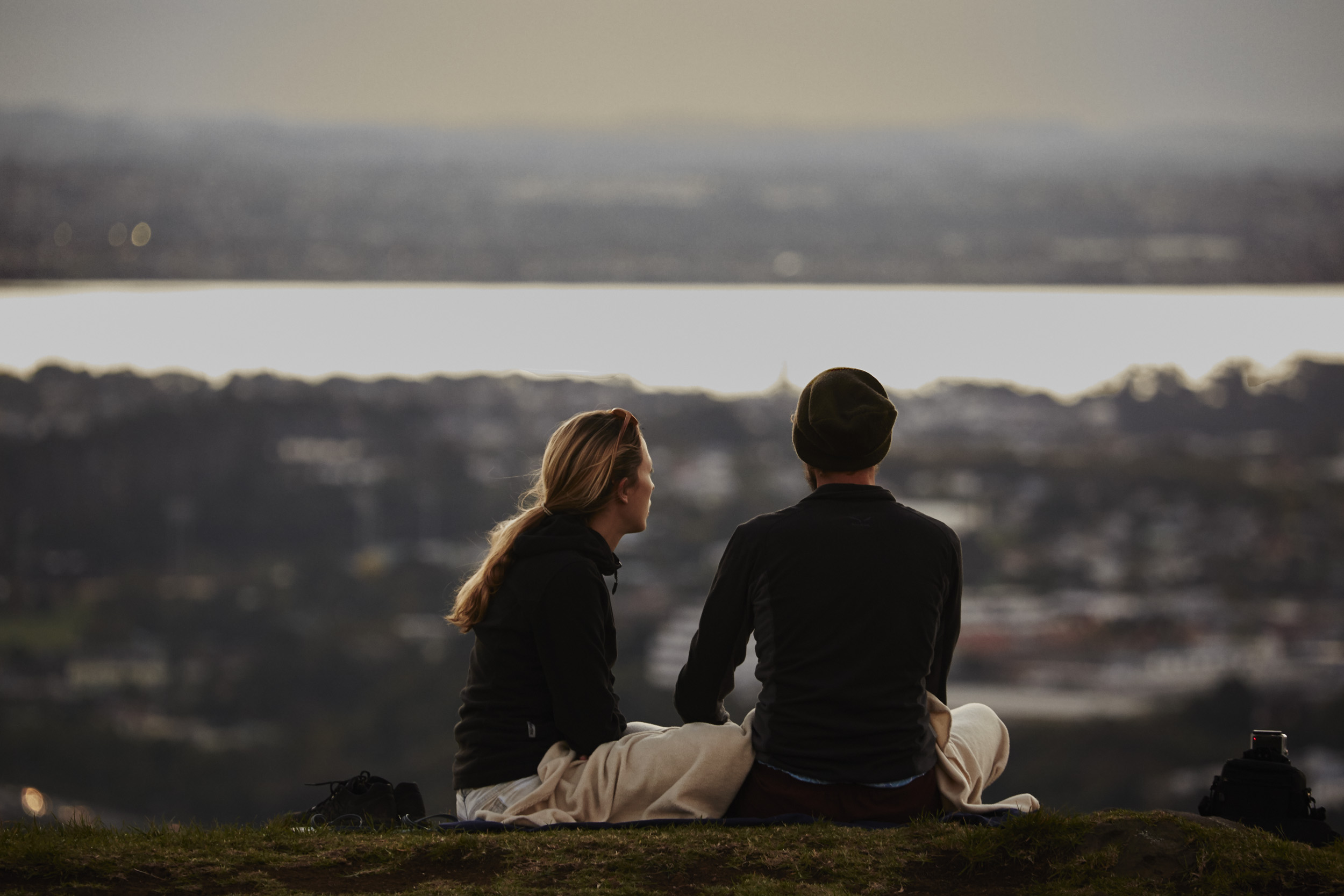 A couple sits on a hill overlooking a lake and town at sunset. The pair is wrapped in a blanket and facing away from the camera.