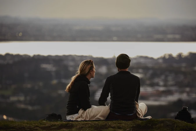 A couple sits on a hill overlooking a lake and town at sunset. The pair is wrapped in a blanket and facing away from the camera.