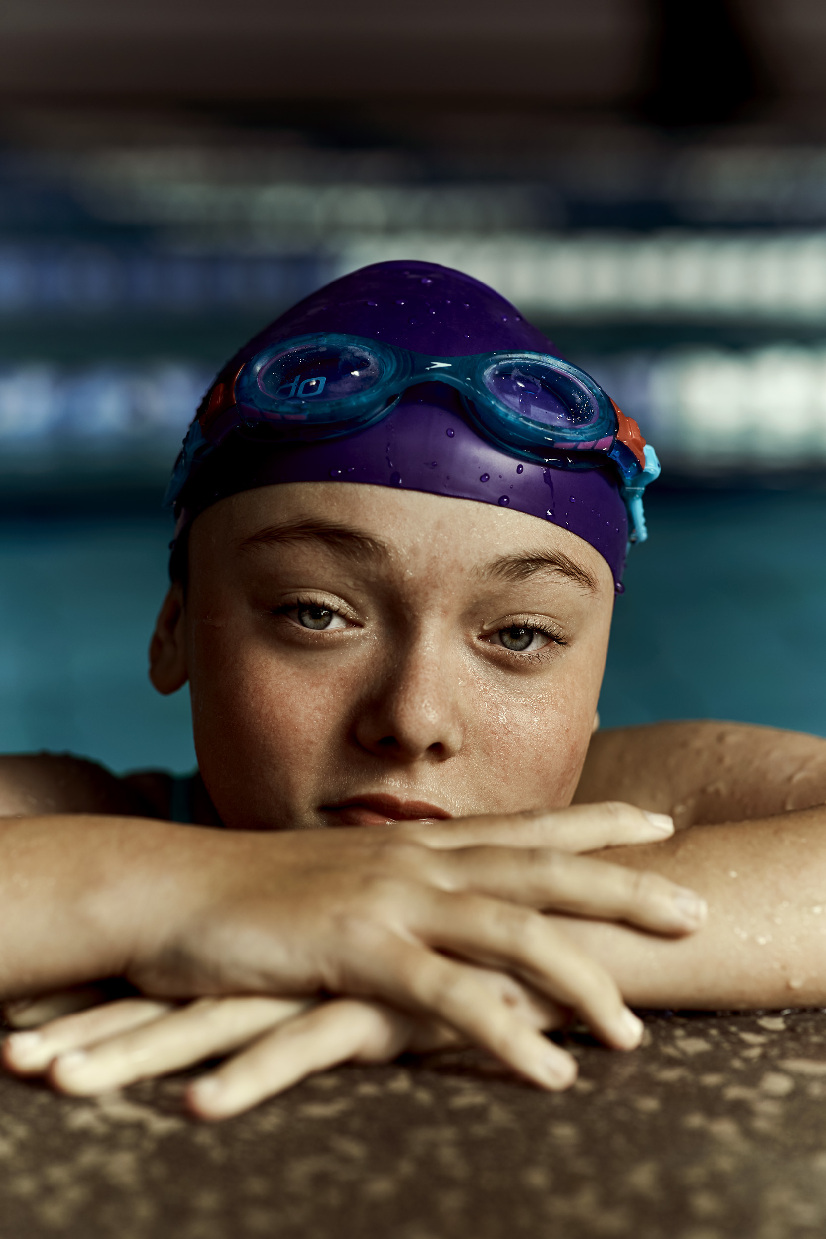 Close-up portrait of a young swimmer with blue goggles and a purple swim cap resting at the edge of an indoor swimming pool.