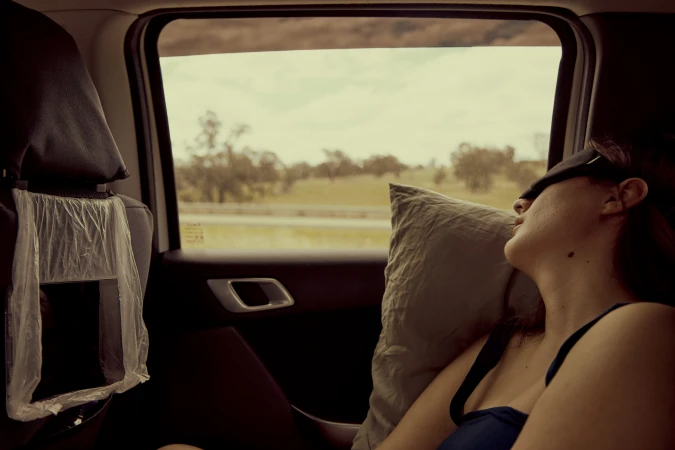 Adult female with a sleep mask dozing with a pillow inside a car, road trip concept with a rural landscape visible through the window.