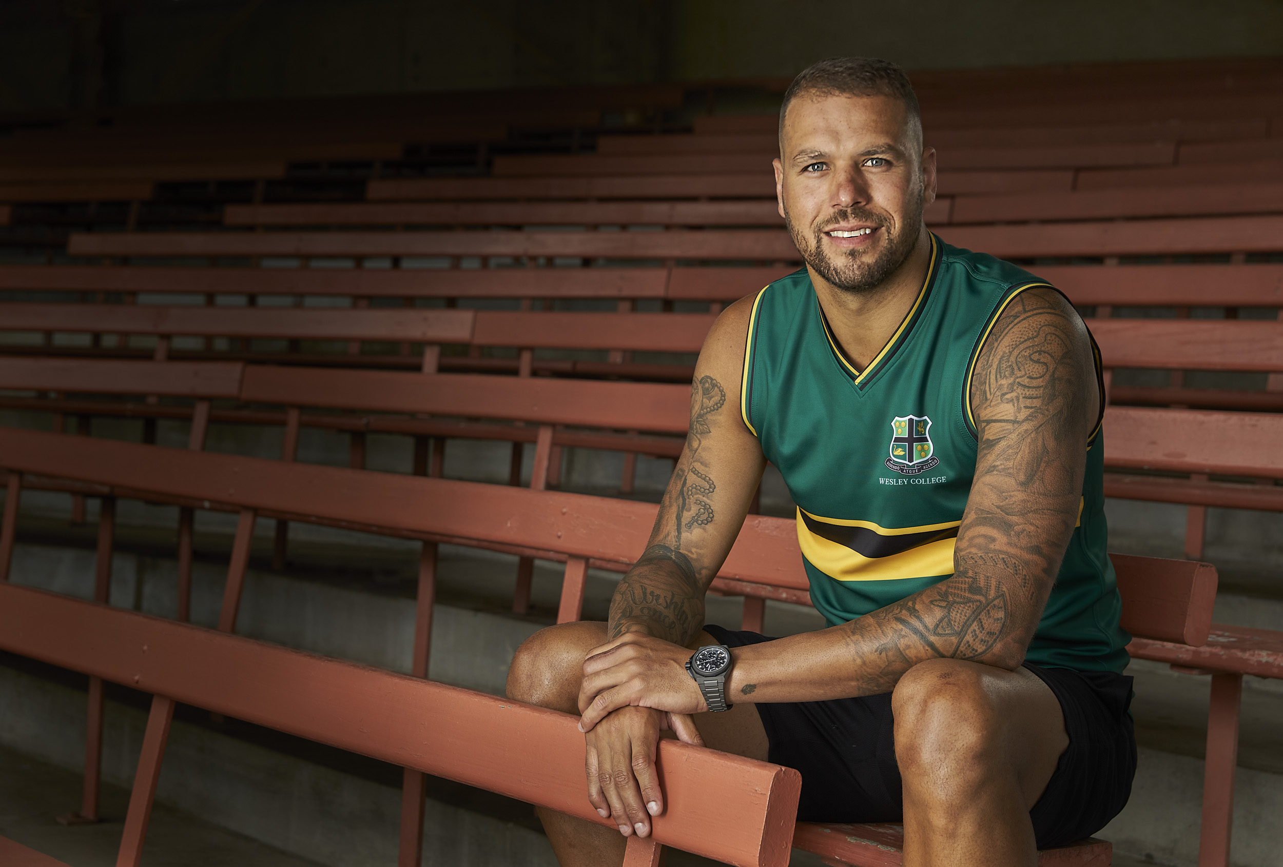 Adult male wearing athletic sleeveless shirt with school logo poses while seated on rows of spectator benches in a covered outdoor setting.