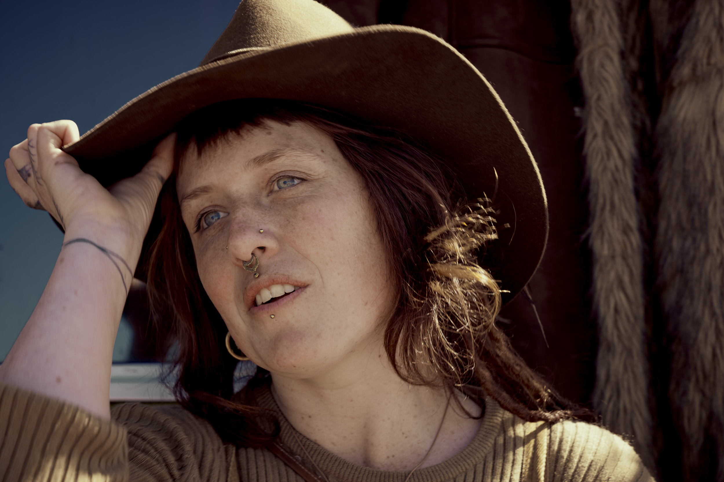Close-up portrait of a woman with freckles, wearing a brown hat and gold earrings, looking upward with a neutral expression.