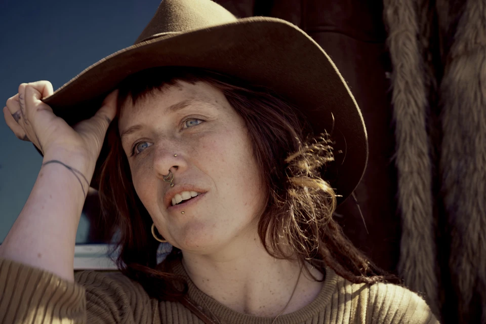 Close-up portrait of a woman with freckles, wearing a brown hat and gold earrings, looking upward with a neutral expression.