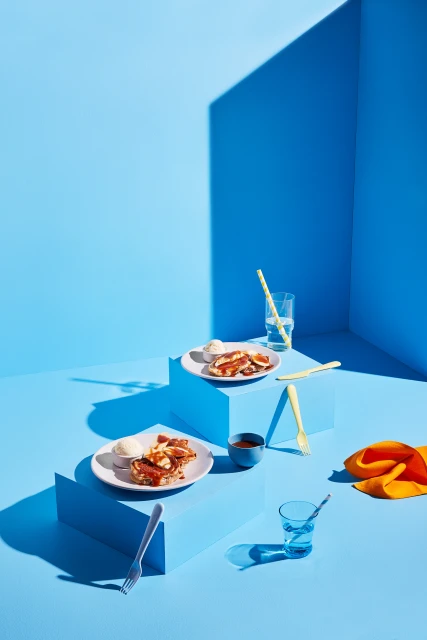Studio shot of stacked blue platforms displaying dessert plates with caramel waffles and ice cream, alongside clear glasses and orange cloth.