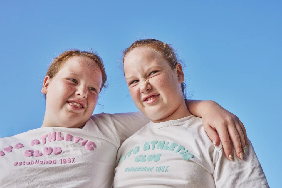 Two smiling, freckled children, one playfully grimacing, pose outdoors under a bright blue sky in matching white shirts.