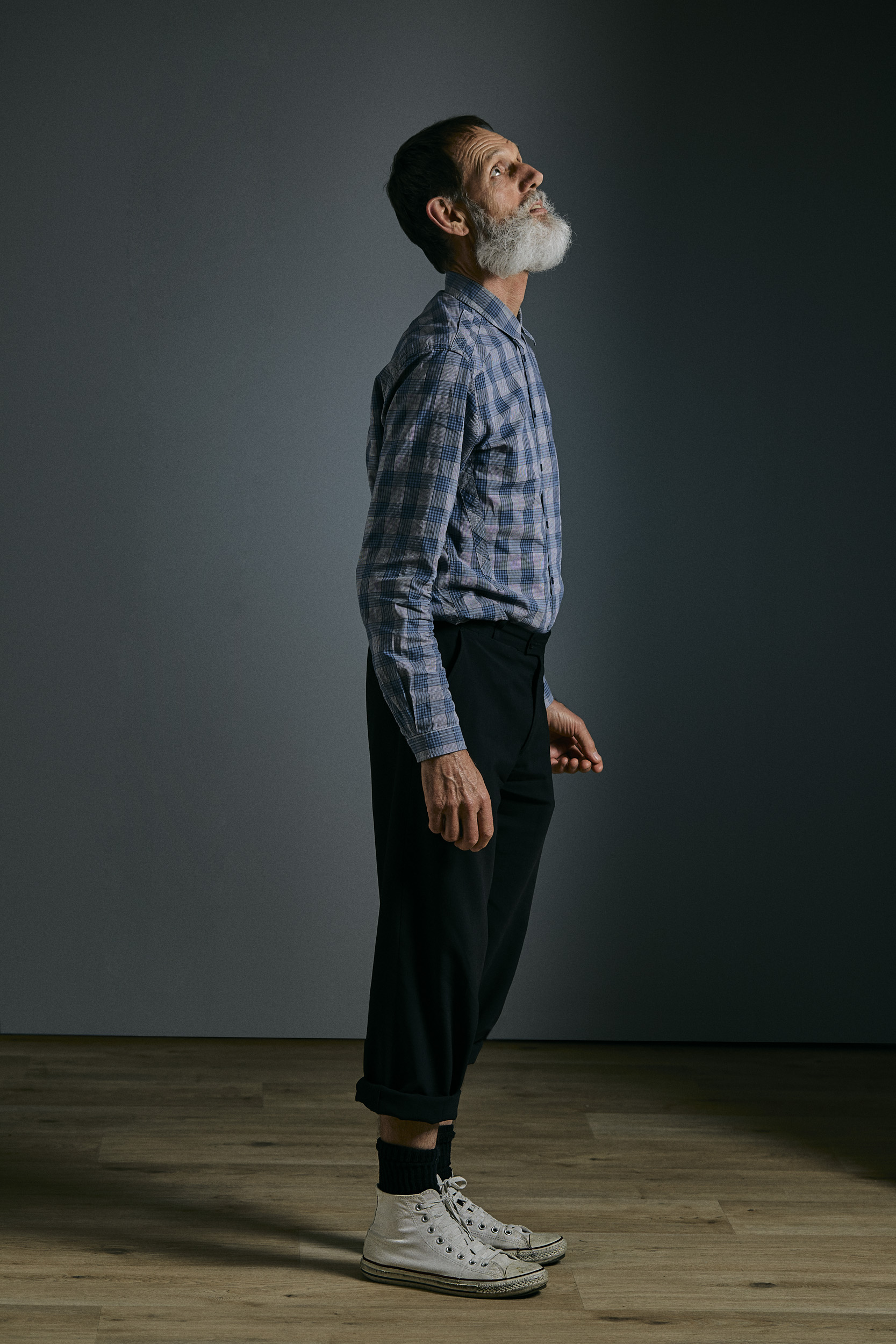 Profile shot of an elderly man with a beard looking up, wearing a plaid shirt, black pants, and white Converse sneakers against grey backdrop.