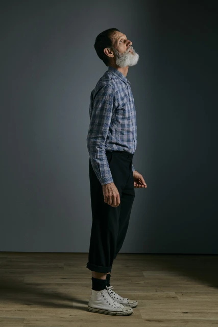 Profile shot of an elderly man with a beard looking up, wearing a plaid shirt, black pants, and white Converse sneakers against grey backdrop.