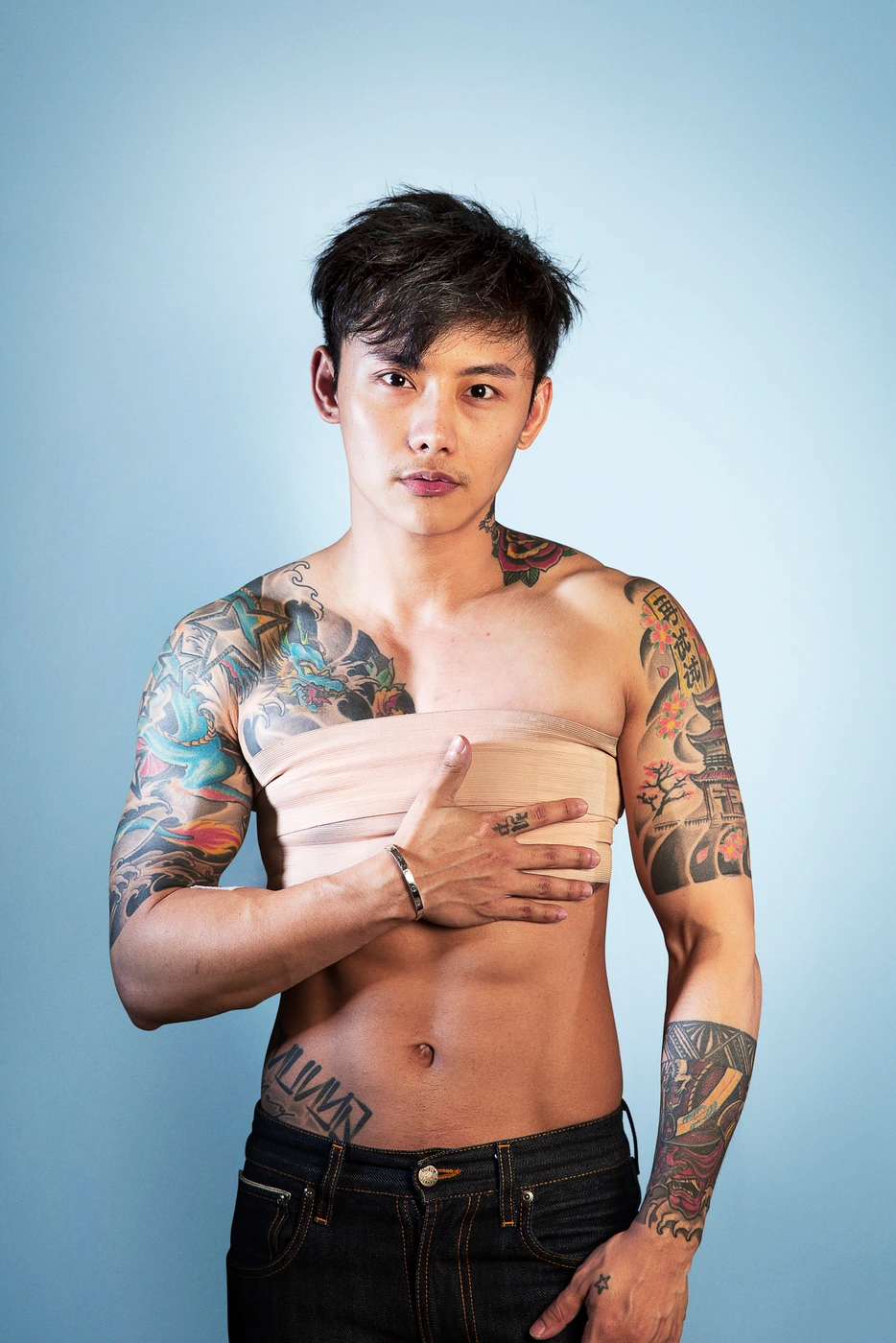 Shirtless man with extensive arm and chest tattoos wearing dark jeans against a light blue studio backdrop