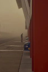 Lone person standing near a pit lane garage beneath a hazy, amber-tinted sky by photographer Sue Stubbs.