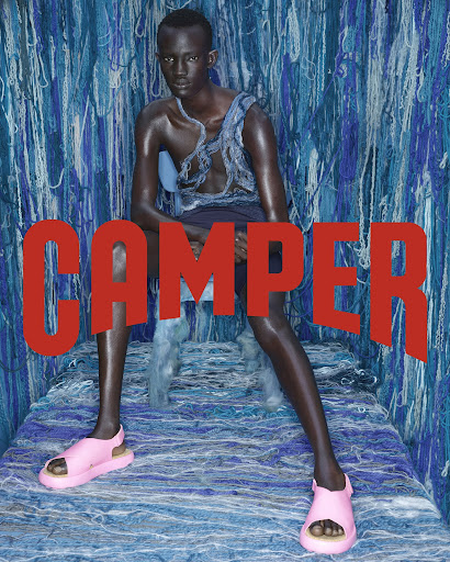 A fashion image featuring a slender, dark-skinned model in sandals, seated against a blue yarn backdrop, with the word CAMPER superimposed in red.