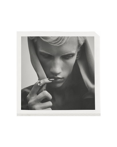 Monochrome portrait of a person with light hair wearing a ring, posed in a square frame against a neutral background.