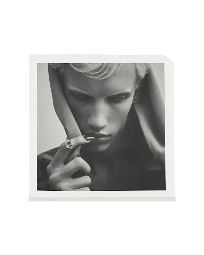 Monochrome portrait of a person with light hair wearing a ring, posed in a square frame against a neutral background.