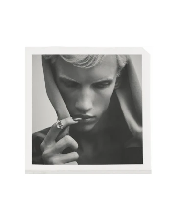 Monochrome portrait of a person with light hair wearing a ring, posed in a square frame against a neutral background.