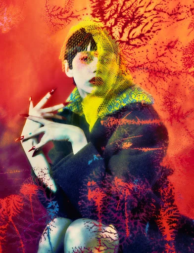 A woman with pale skin, red lipstick, and red nails poses wearing a netted hat and dark coat against a warm orange background.