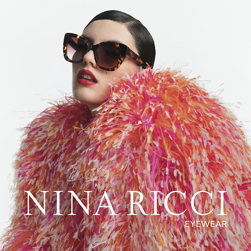 Fashion portrait of a woman wearing tortoiseshell sunglasses and a pink and orange feather coat, posing against a white background.