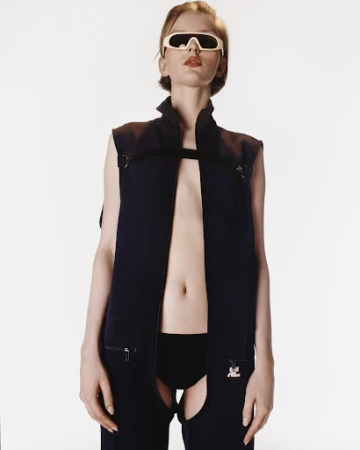 Model wearing dark futuristic sunglasses and a cutout black zip vest against a plain white background