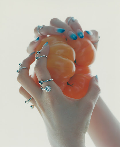 Two hands with blue ombre nails hold an orange pepper against a bright white background. Silver rings adorn each finger.