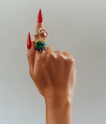 Hand with a brown skin tone pointing up, wearing three large statement rings and red stiletto nails. Plain neutral background.