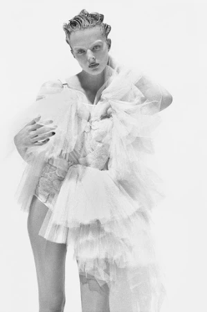 Model with slicked hair wearing voluminous ruffled tulle dress against a plain white studio background