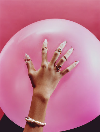 Close-up of a woman's hand adorned with rings and a bracelet, pressing against a pink balloon against a pink backdrop.