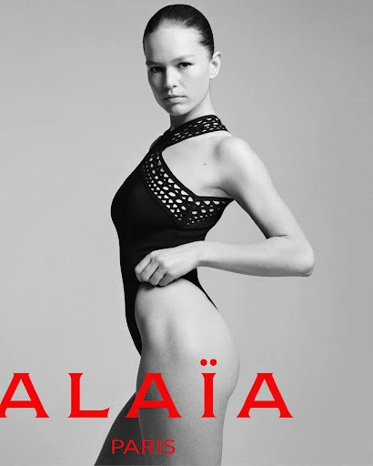 Fashion shot of a young woman posing in a black bodysuit with a high neckline, photographed in a studio with a clean gray background.