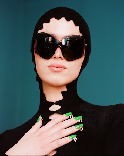 A woman wears dark sunglasses and a black hood with cut-out details, showcasing green nails with lettered charms against a teal background.