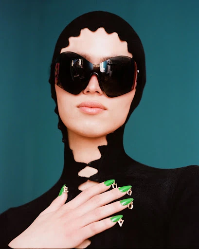 A woman wears dark sunglasses and a black hood with cut-out details, showcasing green nails with lettered charms against a teal background.