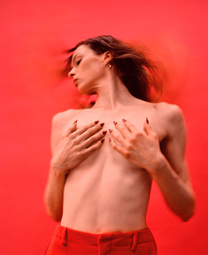 A young adult woman poses with her hands covering her chest against a red backdrop. She has long brown hair and wears red pants.