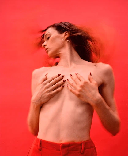 A young adult woman poses with her hands covering her chest against a red backdrop. She has long brown hair and wears red pants.