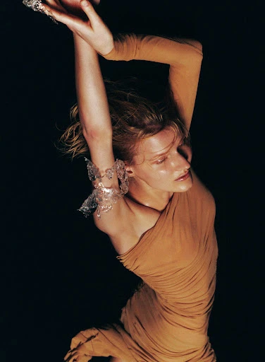 Woman in draped dress with arms up wearing bracelet, against a black backdrop. Fashion editorial style shot with warm lighting.