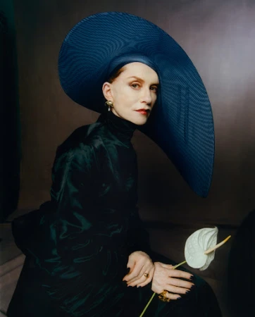 Woman wearing a large wide-brimmed navy blue hat and dark satin dress holding a white anthurium flower