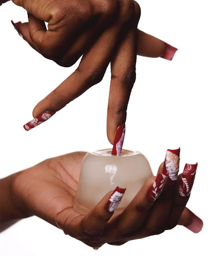 Close-up of dark-skinned hands with decorative nails holding and interacting with a clear, round container against a white background.