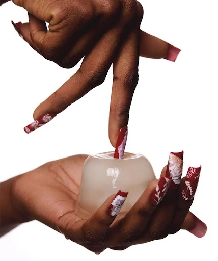 Close-up of dark-skinned hands with decorative nails holding and interacting with a clear, round container against a white background.