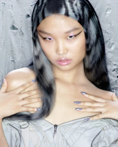 Close-up shot of an Asian woman with grey-streaked hair, dramatic eye makeup, pale pink eyeshadow, and a grey top, posing against a textured grey background.