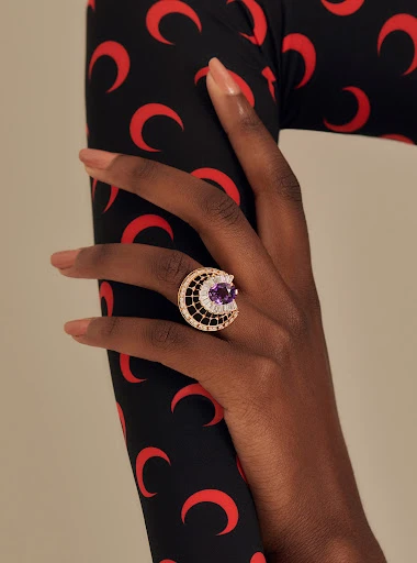 Close-up of a dark-skinned hand with tan painted nails wearing a ring with a purple gemstone and black and gold setting, against a fabric background.