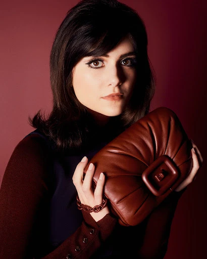 Portrait of a dark-haired woman holding a brown leather bag and wearing a brown bracelet and long-sleeved brown and blue dress.