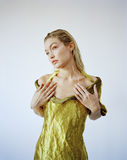 Gigi Hadid wearing an olive green satin dress with wet-look blonde hair against a plain white studio background