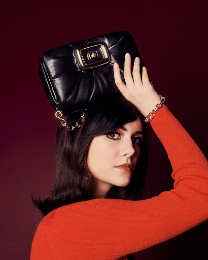 A woman with dark hair holds a black leather handbag with gold hardware above her head while posing against a dark red background.