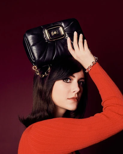 A woman with dark hair holds a black leather handbag with gold hardware above her head while posing against a dark red background.