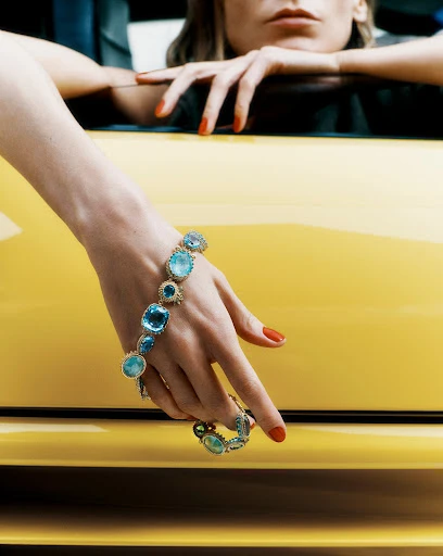 Woman's hand with red nails displays a blue gemstone bracelet on a yellow car; the image is a close-up with a shallow depth of field.