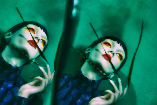 Double exposure portrait of a woman with stylized makeup in blue top, geometric earrings, and dark sprigs against a teal background.