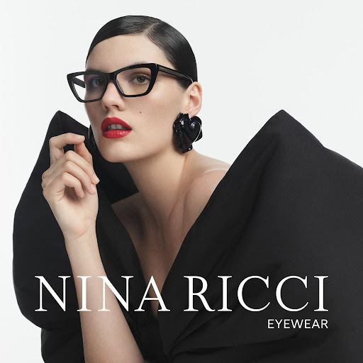 A woman with dark hair and red lipstick models black eyeglasses, wearing a black dress and black earrings against a white backdrop.